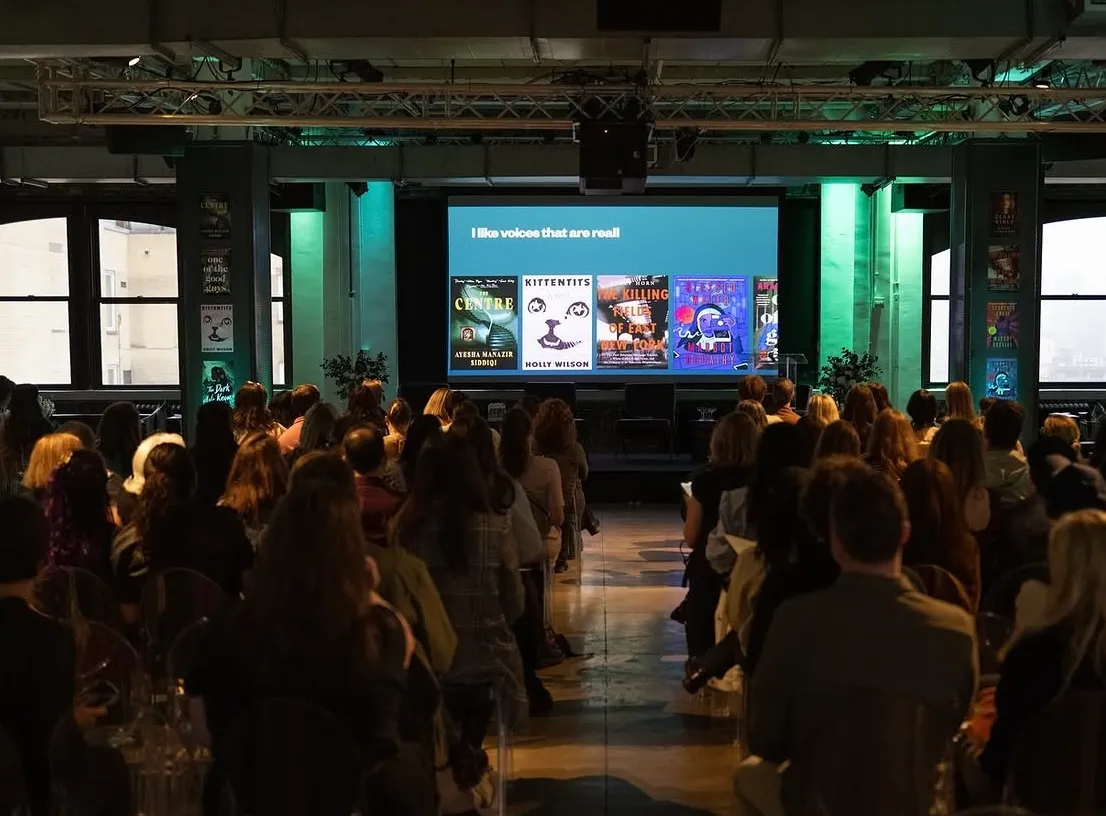 Darkened room full of event attendees from behind viewing a screen on stage with books on the slide.