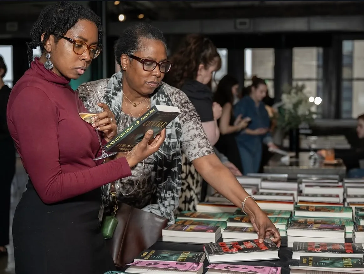 Two women browsing a table full of books for sale on it. One woman is reading the back of a books content as she holds her wine glass to her chest. The other woman reaches for a book in the middle row on the table.