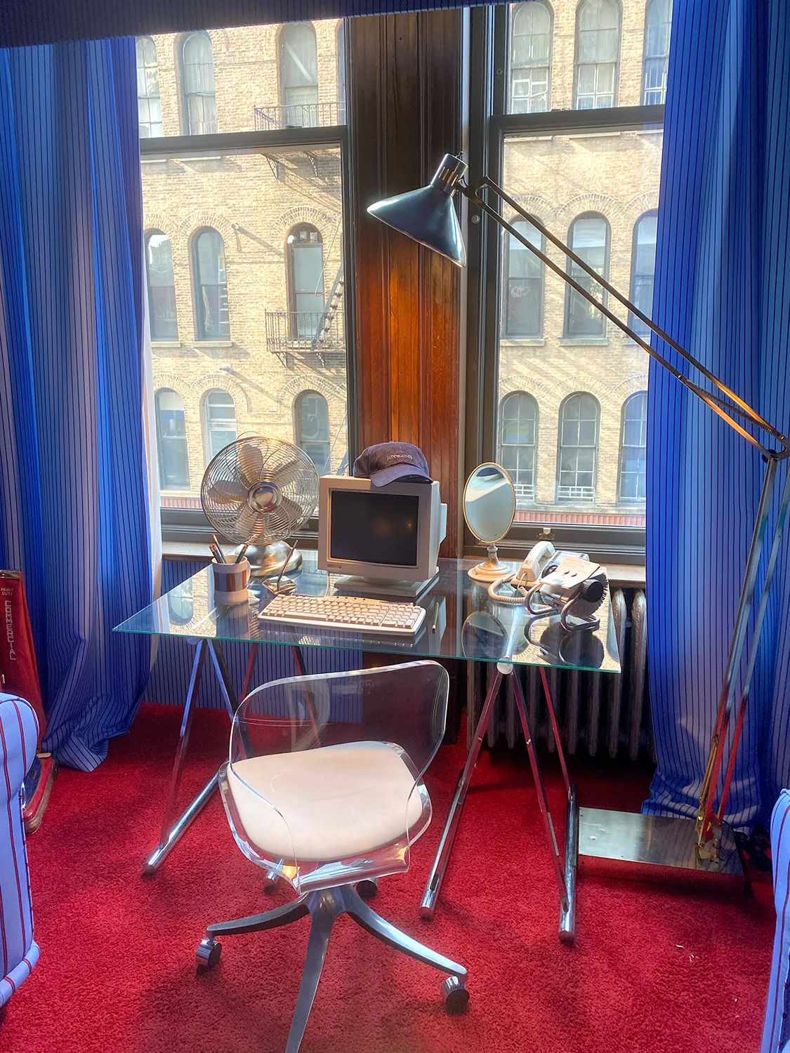 Glass and metal office desk with vintage computer and other objects on it in room with clear plastic rolling office chair, facing big windows, red carpet and blue striped curtains