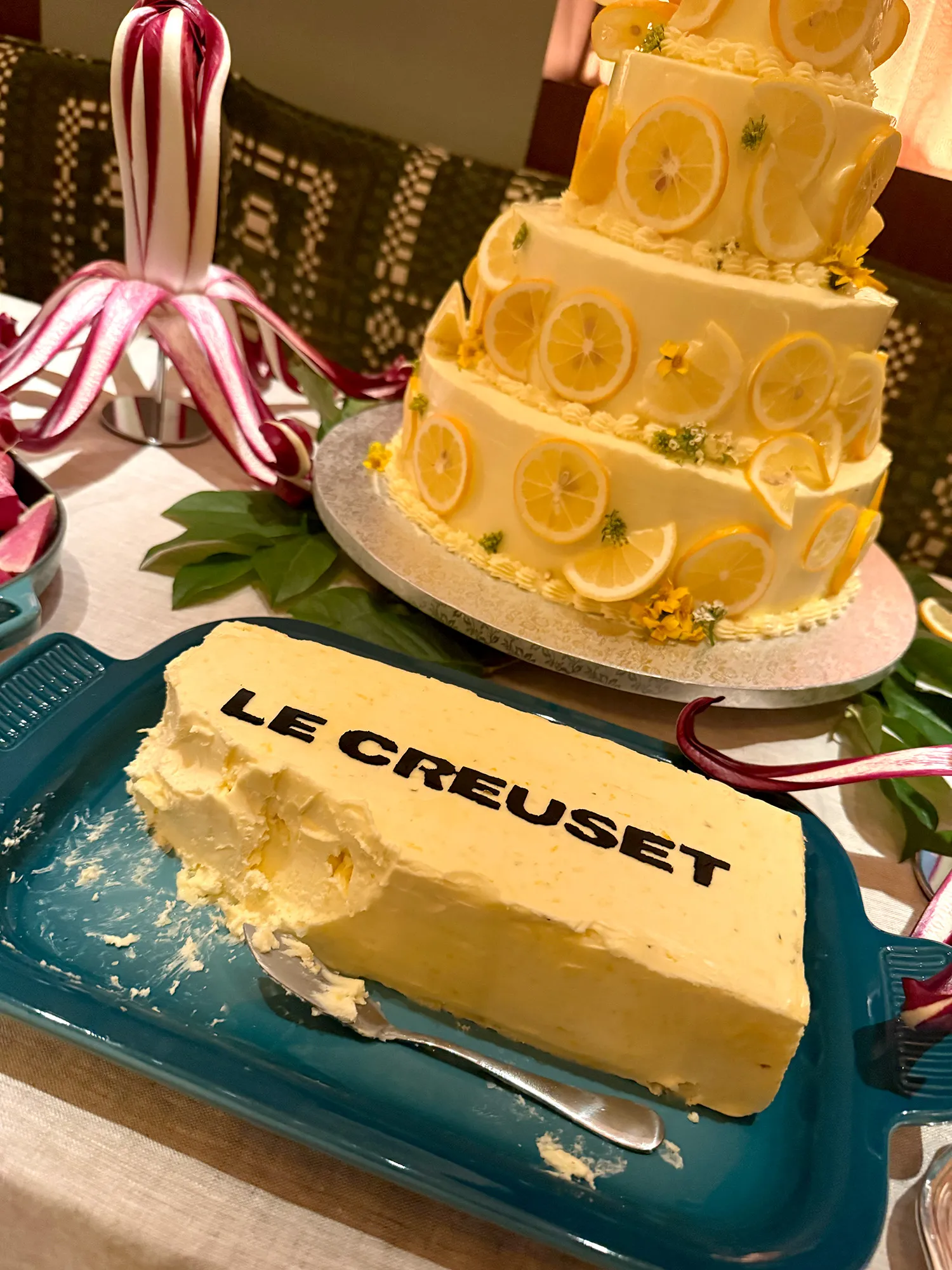 Light yellow frosted cake with the words Le Creuset on it and a 4 tiered, frosted cake decorated with lemons in the background