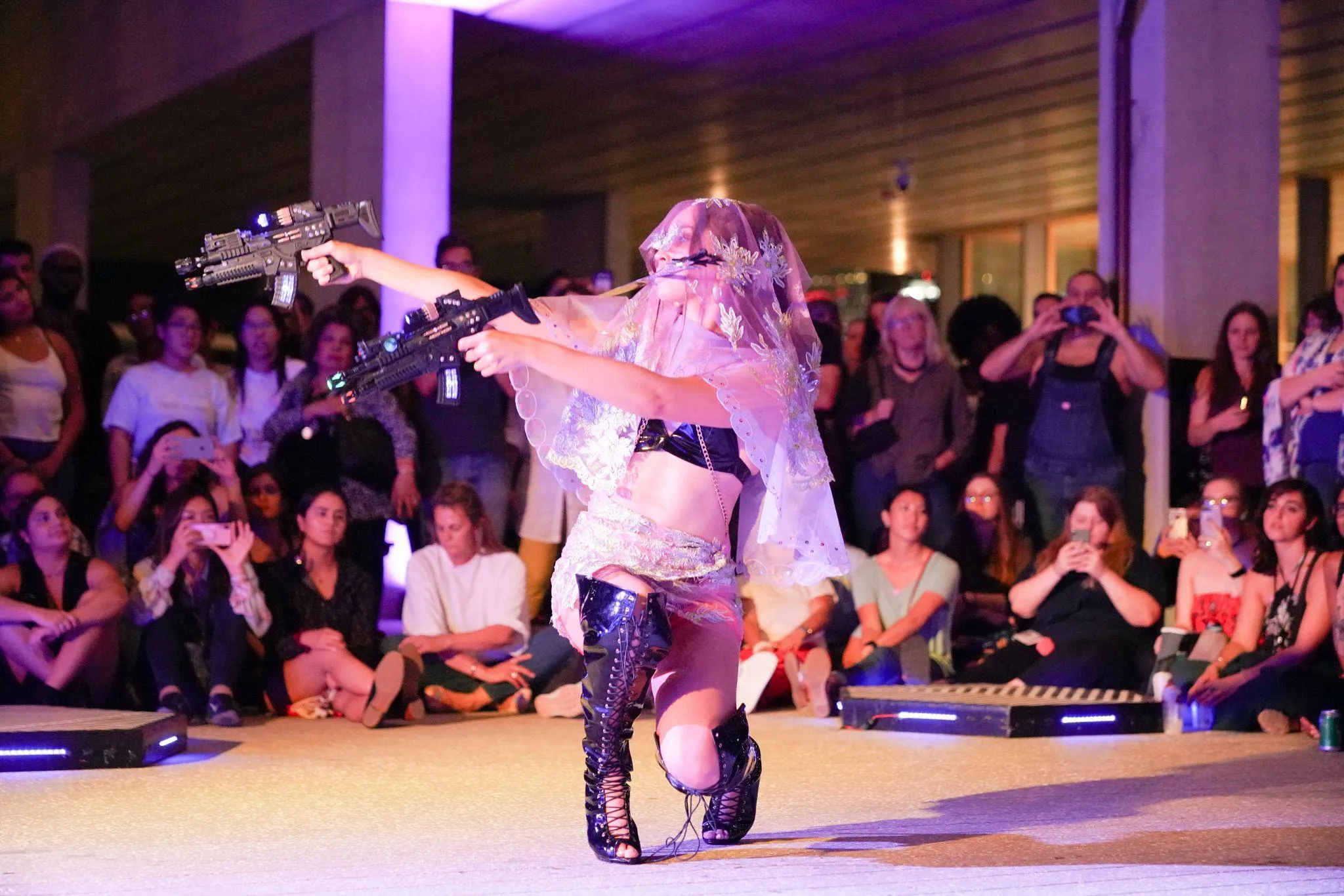 Performer in veiled and vinyl costume points two toy machine guns as they crouch down