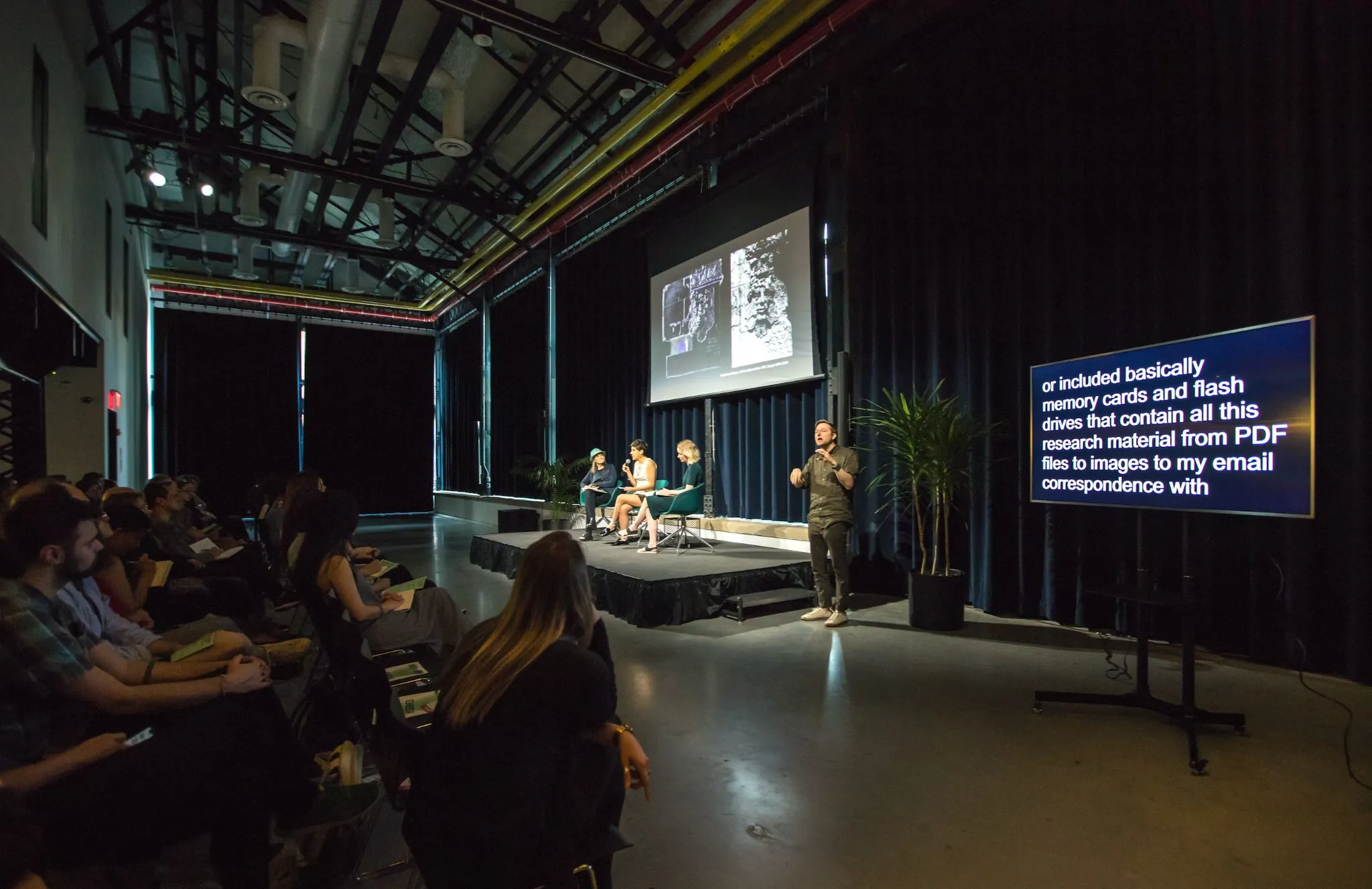 Stage with 3 panelists and projection screen while audience listens, captions and sign language are offered